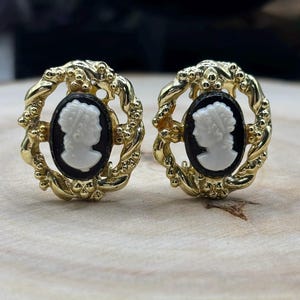Peut inclure: Une paire de boucles d'oreilles ovales présentant un camée blanc représentant un profil de personne sur fond noir. Le camée est entouré d'une monture en métal doré texturé avec un motif décoratif en forme de corde torsadée.