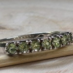 May include: A silver-toned ring featuring a row of six small, round, light green gemstones set in a prong setting. The ring is positioned alongside a thin wooden dowel, highlighting the band's metallic finish and the faceted cut of the stones.
