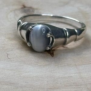 Op de afbeelding: Een zilveren ring met een gladde, ovaalvormige, lichtgrijze cabochon edelsteen in een zetting met vier pootjes. De ringband heeft een gepolijste metalen afwerking met een gesegmenteerd, afgerond ontwerp dat naar achteren toe taps toeloopt.