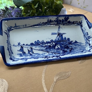 May include: A rectangular ceramic serving tray featuring a blue and white transferware pattern. The design depicts a Dutch landscape with a prominent windmill, sailboats on water, and houses. The tray has integrated handles and a blue border.