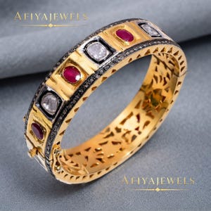 May include: A gold hinged bangle bracelet featuring alternating oval-shaped red rubies and clear white stones set in dark metal frames. The outer band displays a geometric pattern with small black stones, while the inner band has a cut-out design.