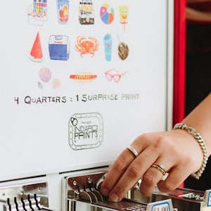 May include: A white mini print vending machine with red trim, featuring colourful beach-themed illustrations and the text "MINI PRINT VENDING MACHINE" and "4 QUARTERS = 1 SURPRISE PRINT". The machine has metal coin slots labelled $1.00.