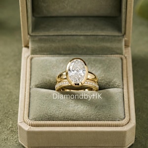 Puede incluir: Un conjunto de anillos de oro con una piedra central de corte ovalado en engaste de bisel, junto a una alianza a juego con pequeños diamantes pavé. La joya se presenta en un estuche forrado de terciopelo verde salvia con el texto DiamondbyHK.