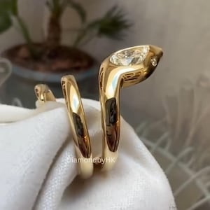May include: A gold snake-shaped ring featuring a smooth, polished finish and a pear-shaped diamond set in the head. The design wraps around the finger, and the text DiamondbyHK is visible on the side of the band. The metal is reflective and bright.