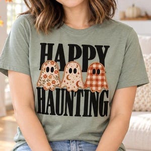 Peut inclure: Un t-shirt à manches courtes vert sauge avec le texte noir "HAPPY HAUNTING" et trois illustrations de fantômes. Les fantômes présentent des motifs différents : un avec des fleurs orange, un avec des lunes et des étoiles, et un à carreaux orange et blanc.