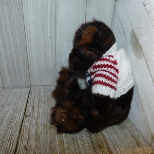 May include: A brown teddy bear wearing a white and red striped sweater. The bear is sitting on a wooden surface.