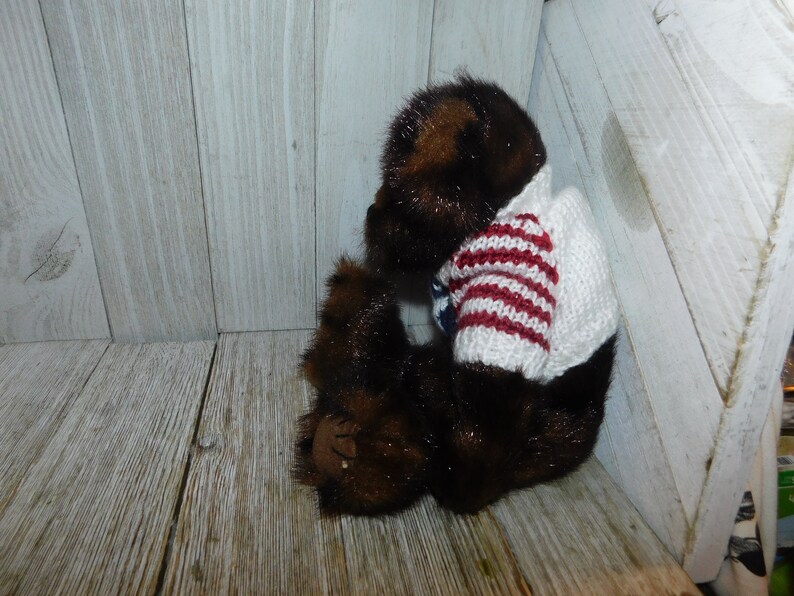 May include: A brown teddy bear wearing a white and red striped sweater. The bear is sitting on a wooden surface.