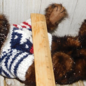 May include: A brown and black plush toy wearing a white, blue and red knitted sweater. The toy is lying on a wooden surface.