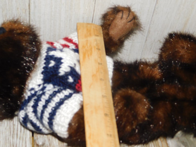 May include: A brown and black plush toy wearing a white, blue and red knitted sweater. The toy is lying on a wooden surface.