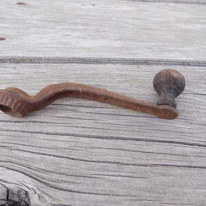 May include: A vintage metal hand crank featuring a rusted, weathered finish. The crank has a curved arm, a circular gear-like attachment at one end, and a small, rounded wooden knob at the opposite end for manual operation and rotation.