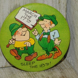 Puede incluir: Un bot&oacute;n verde y amarillo con dos personajes de dibujos animados. Los personajes llevan sombreros verdes y sostienen carteles. El cartel de la izquierda dice "March the 17th" y el cartel de la derecha dice "Sleep the 18th!"