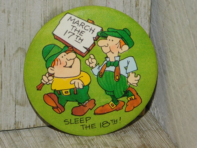 Puede incluir: Un bot&oacute;n verde y amarillo con dos personajes de dibujos animados. Los personajes llevan sombreros verdes y sostienen carteles. El cartel de la izquierda dice "March the 17th" y el cartel de la derecha dice "Sleep the 18th!"