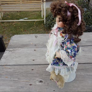 May include: A vintage porcelain doll with brown curly hair, a pink ribbon, and a floral dress. The doll has a white lace dress and a pearl necklace. It is standing on a white stand. The doll is approximately 12 inches tall.