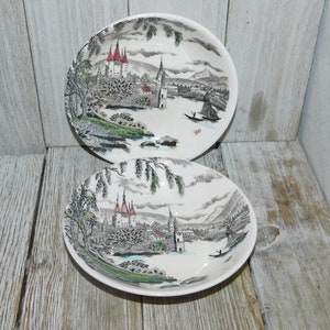 May include: Two white ceramic bowls with a black and white landscape design featuring a castle with red roofs and a boat on a lake. The bowls are on a light wood surface.
