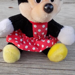 May include: A plush Minnie Mouse doll, featuring a black body, white gloves, and yellow feet. The doll wears a red dress with white polka dots and a red collar. The doll has a beige face with black eyes and a black nose.