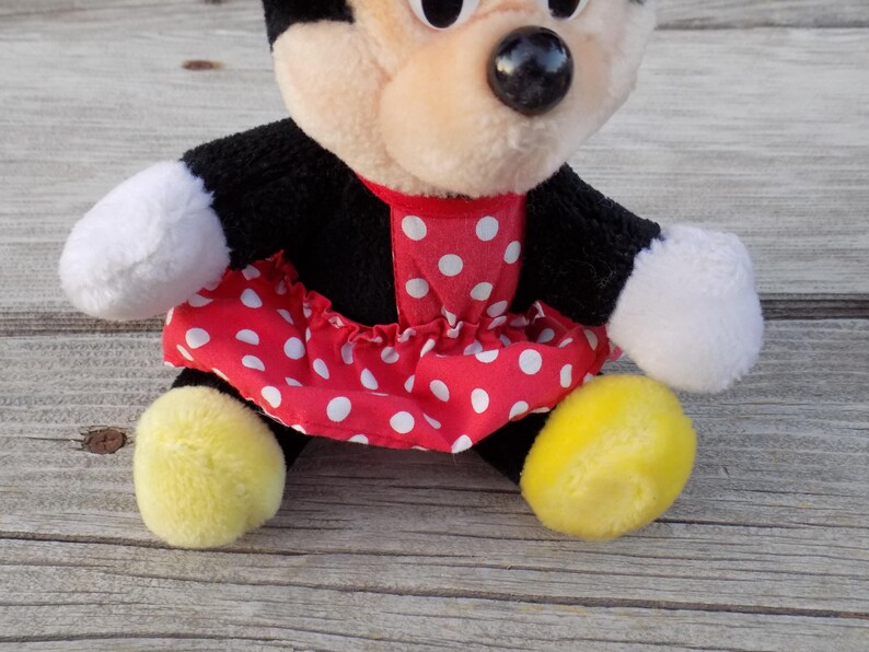 May include: A plush Minnie Mouse doll, featuring a black body, white gloves, and yellow feet. The doll wears a red dress with white polka dots and a red collar. The doll has a beige face with black eyes and a black nose.