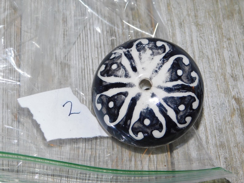 K&ouml;nnte beinhalten: Ein runder Keramikknopf mit einem wei&szlig;en und schwarzen Blumendesign. Der Knopf hat ein Loch in der Mitte.