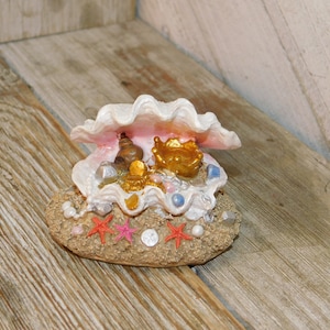 May include: A decorative seashell figurine with a treasure chest theme. The open shell reveals gold coins, a crown, and a small bottle. The base is sandy with starfish and pearls. The shell is white and pink.