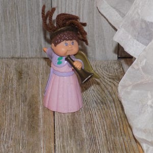 May include: A Cabbage Patch Kid angel doll with brown hair, wearing a pink dress and holding a gold trumpet.