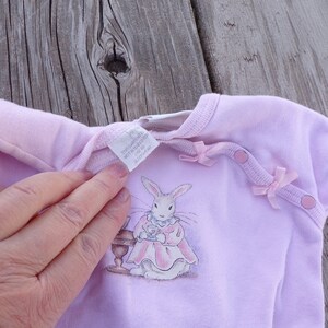 May include: A pale pink baby onesie with a white bunny rabbit illustration wearing a pink dress and holding a teacup. The onesie has two pink bows and two pink buttons on the shoulders. The tag reads "The Launderette, Not Ironed, 100% Cotton, Size 0-3 Months, 012779148".