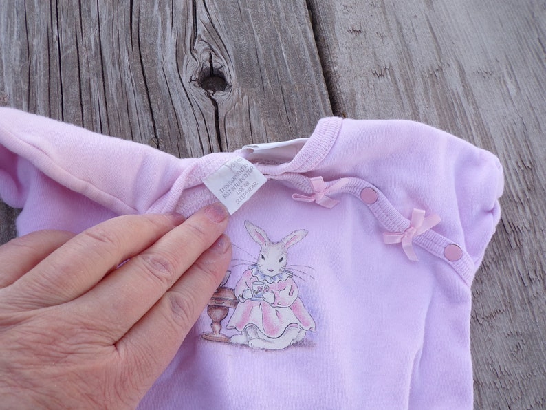 May include: A pale pink baby onesie with a white bunny rabbit illustration wearing a pink dress and holding a teacup. The onesie has two pink bows and two pink buttons on the shoulders. The tag reads "The Launderette, Not Ironed, 100% Cotton, Size 0-3 Months, 012779148".