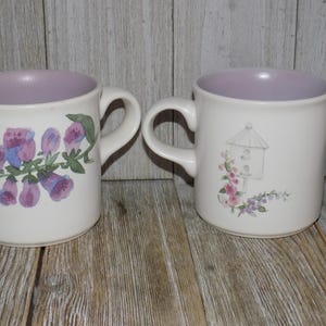 May include: Two white ceramic mugs with lavender interiors. One mug features a floral design with purple and blue flowers, the other has a birdhouse and floral accents. Both mugs have curved handles.