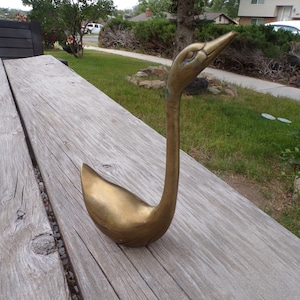 Pu&ograve; includere: Una statuetta di cigno in metallo dorato con un collo lungo e un corpo curvo. Il cigno &egrave; seduto su una superficie di legno.