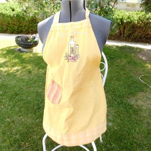 May include: Yellow kitchen apron with a gingham border and a striped pocket. The apron has an embroidered design of bottles and the text "Good  Appetite".
