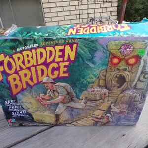 May include: A board game box with a green, yellow, and purple design. The game is called "Forbidden Bridge" and features a motorized adventure game. The box shows a jungle scene with a stone idol with glowing red eyes and a bridge.