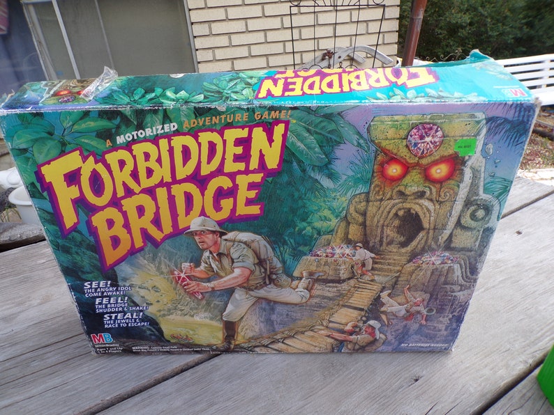 May include: A board game box with a green, yellow, and purple design. The game is called "Forbidden Bridge" and features a motorized adventure game. The box shows a jungle scene with a stone idol with glowing red eyes and a bridge.