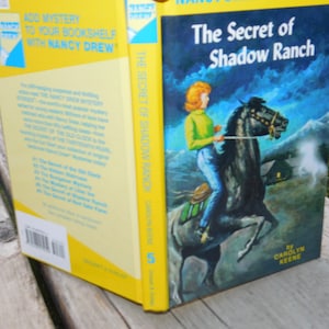 May include: A yellow and blue Nancy Drew book cover with the title "The Secret of Shadow Ranch" and an illustration of a young woman riding a black horse. The book is number 5 in the series.
