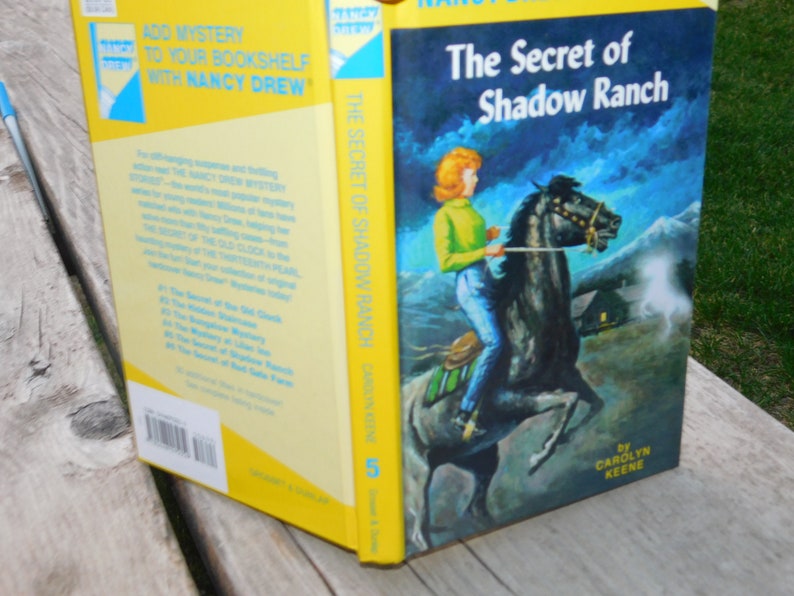 May include: A yellow and blue Nancy Drew book cover with the title "The Secret of Shadow Ranch" and an illustration of a young woman riding a black horse. The book is number 5 in the series.