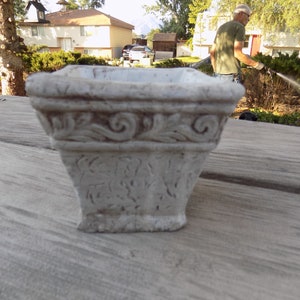 May include: A white ceramic planter with an ornate design around the top edge. The planter is square shaped and has a slightly flared opening.