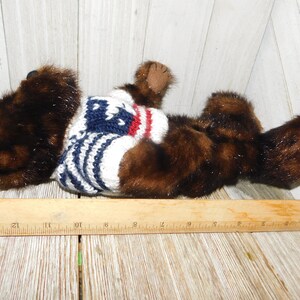 May include: A brown and black plush bear wearing a white, blue and red striped sweater. The bear is lying on its side with its head turned to the left.