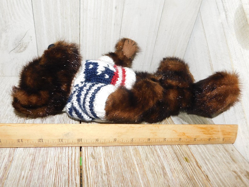 May include: A brown and black plush bear wearing a white, blue and red striped sweater. The bear is lying on its side with its head turned to the left.