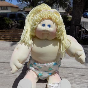 May include: A vintage doll with a soft, cream-colored body, light yellow yarn hair, and blue eyes. The doll wears a diaper with a pattern of frogs and other designs. The doll is sitting on a wooden surface.