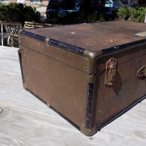 May include: A vintage brown trunk with black trim and brass hardware. The trunk has metal corner protectors and a handle. The trunk is likely used for storage or travel.