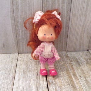 Pode incluir: Uma boneca de cabelo ruivo com uma camisa xadrez rosa e branca, cal&ccedil;as rosa e sapatos rosa. A boneca tem um la&ccedil;o vermelho no cabelo e uma express&atilde;o doce no rosto.