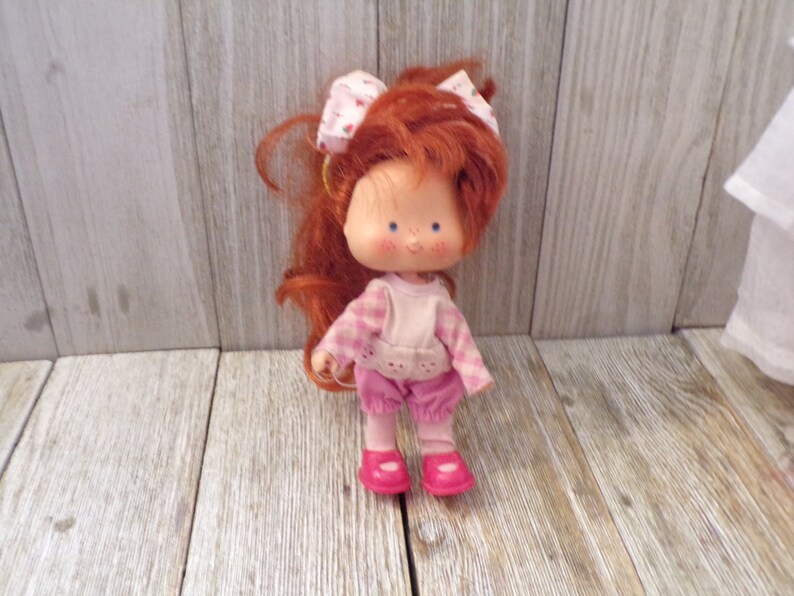 Pode incluir: Uma boneca de cabelo ruivo com uma camisa xadrez rosa e branca, cal&ccedil;as rosa e sapatos rosa. A boneca tem um la&ccedil;o vermelho no cabelo e uma express&atilde;o doce no rosto.