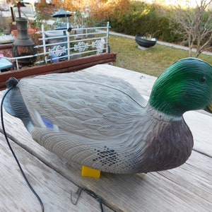 May include: A realistic-looking decoy duck with a green head and a gray body. The duck is made of plastic and has a string attached to it.
