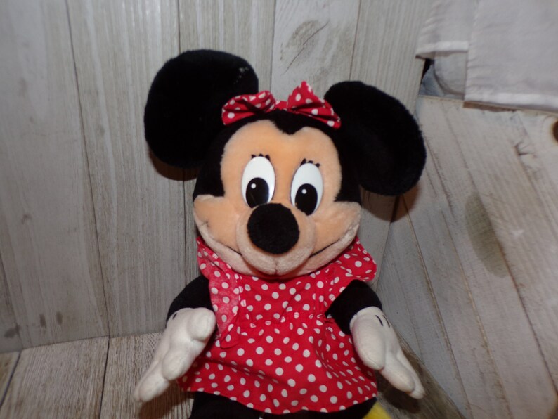 K&ouml;nnte beinhalten: Ein Pl&uuml;schtier von Minnie Mouse, das ein rotes Kleid mit wei&szlig;en Punkten und eine rote Schleife tr&auml;gt. Das Spielzeug hat schwarze Ohren und eine schwarze Nase.