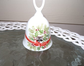 May include: White porcelain Christmas bell with a red ribbon and holly leaves. A raccoon's face is peeking out from behind the holly.