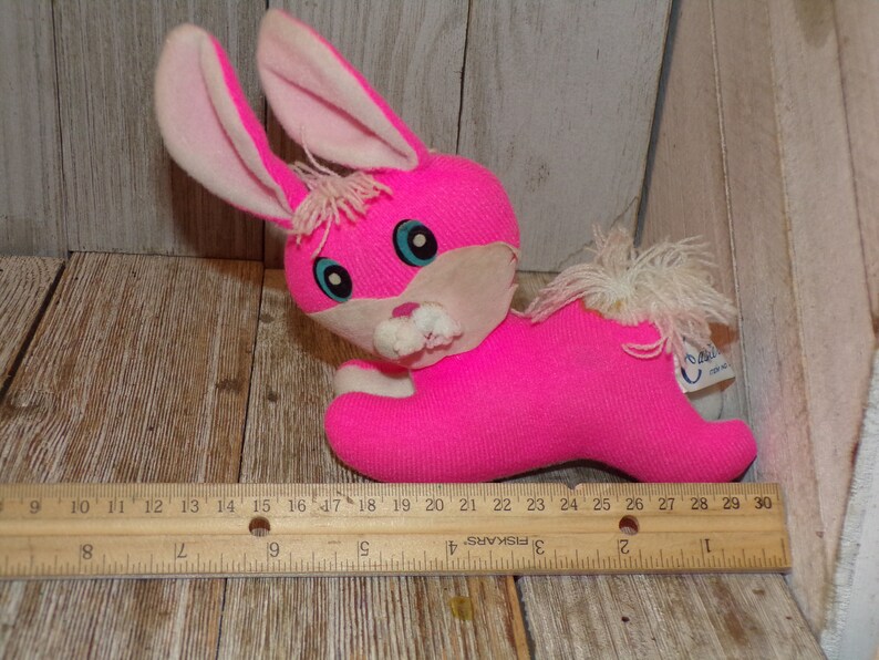 May include: Pink plush bunny rabbit toy with white ears and a white fluffy tail. The bunny has blue eyes and a white nose and mouth. The toy is approximately 10 inches long.