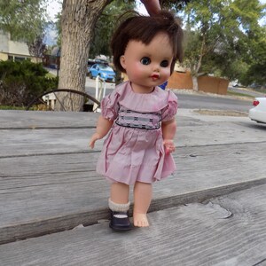 May include: A doll with short brown hair and blue eyes wearing a light pink dress with a ruffled collar and smocked bodice detail. The doll has one dark shoe with a white sock on its right foot and is bare on its left foot, featuring painted lips.