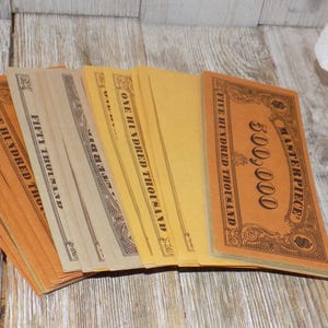 May include: A collection of vintage-style play money in various colors, including orange, tan, and yellow. The bills are labeled with denominations like "Five Hundred Thousand" and "Fifty Thousand". The words "Masterpiece" are also visible.