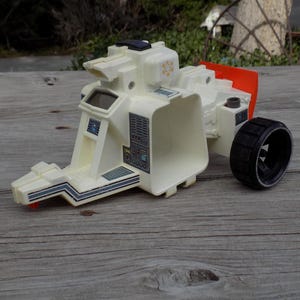 May include: A vintage off-white toy vehicle with a black wheel and orange accents. The toy has a futuristic design with various panels, a screen, and a detailed front section. It is set on a weathered wooden surface.