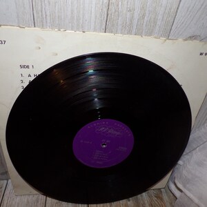 Puede incluir: Un disco de vinilo negro con una etiqueta morada que dice "Alshire Presents Etta James" y "At Last". El disco est&aacute; en una funda de papel blanco con el texto "W 9037" impreso en la esquina superior derecha y "Side 1" impreso en el lado izquierdo.