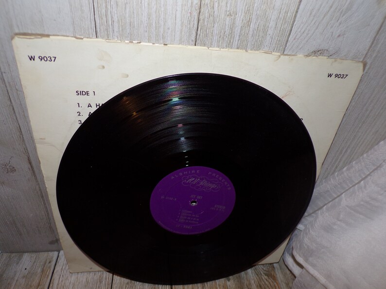 Puede incluir: Un disco de vinilo negro con una etiqueta morada que dice "Alshire Presents Etta James" y "At Last". El disco est&aacute; en una funda de papel blanco con el texto "W 9037" impreso en la esquina superior derecha y "Side 1" impreso en el lado izquierdo.
