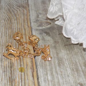 May include: A gold charm bracelet with a rocking horse, bell, and other decorative charms. The bracelet is on a wooden surface, with white lace fabric in the background. The bracelet's design is intricate and detailed.
