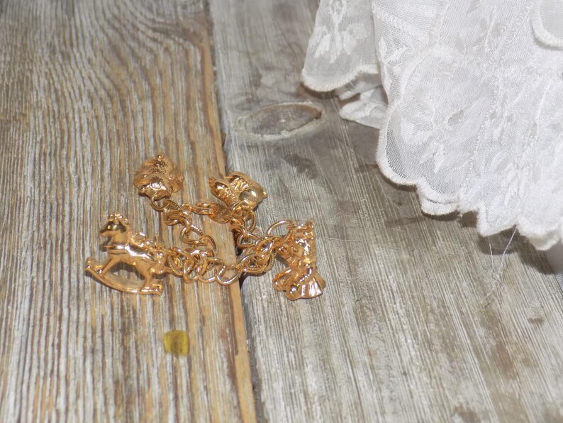 May include: A gold charm bracelet with a rocking horse, bell, and other decorative charms. The bracelet is on a wooden surface, with white lace fabric in the background. The bracelet's design is intricate and detailed.